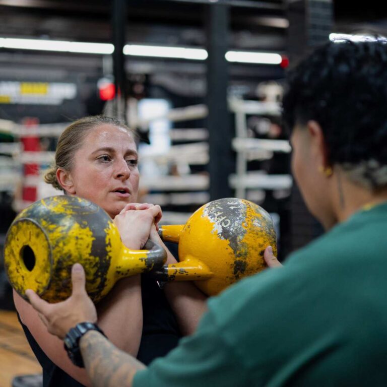 Women lifting 2 yellow kettlebells and coach correcting it